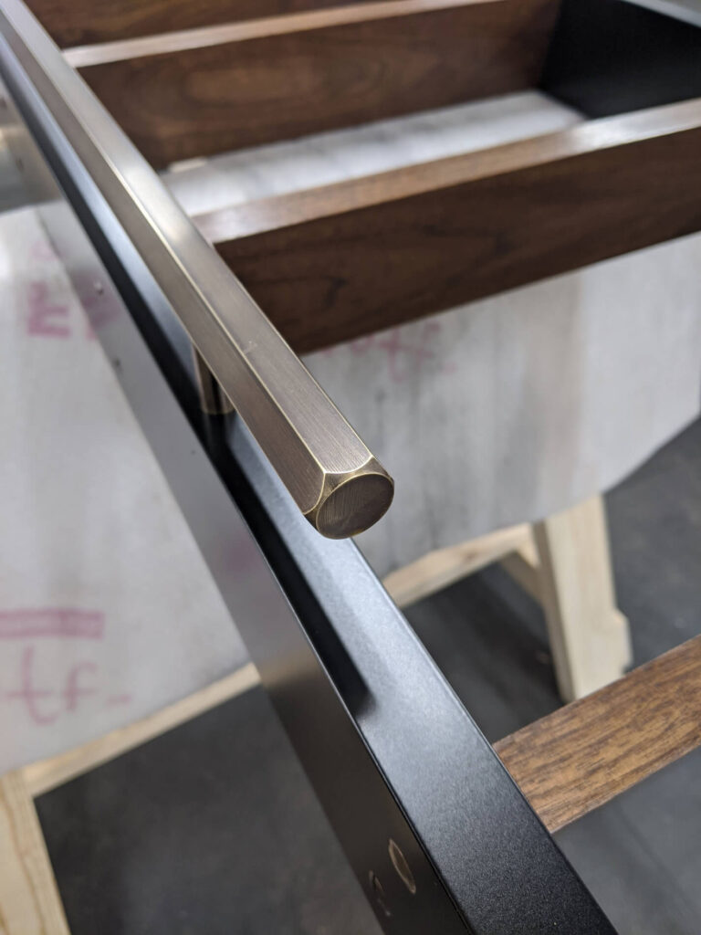 Close-up of a modern furniture piece showing a brushed metal handle attached to a black frame with wooden panels, set against a blurred workshop background.