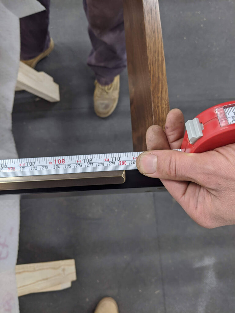 A person measures a wooden edge with a tape measure, showing a measurement of about 33 inches. Part of two people’s legs and shoes are visible in the background on a dark floor.