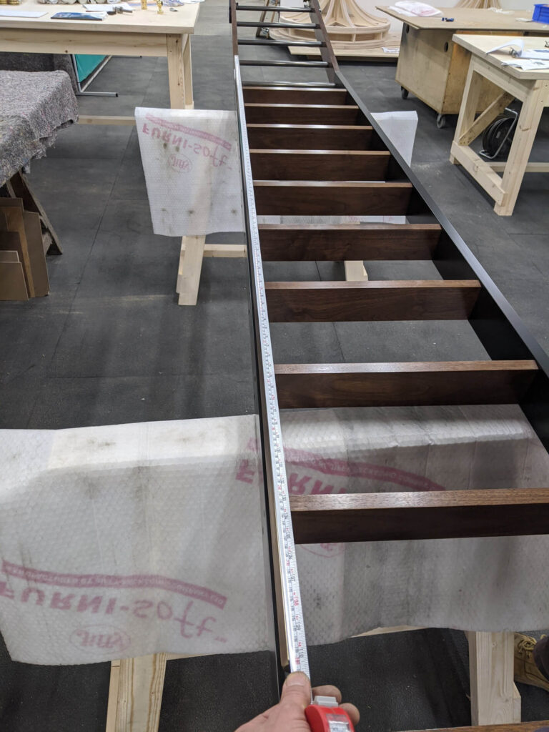 A person holds a measuring tape along the side of a wooden staircase frame in a workshop, with workbenches and tools visible in the background.