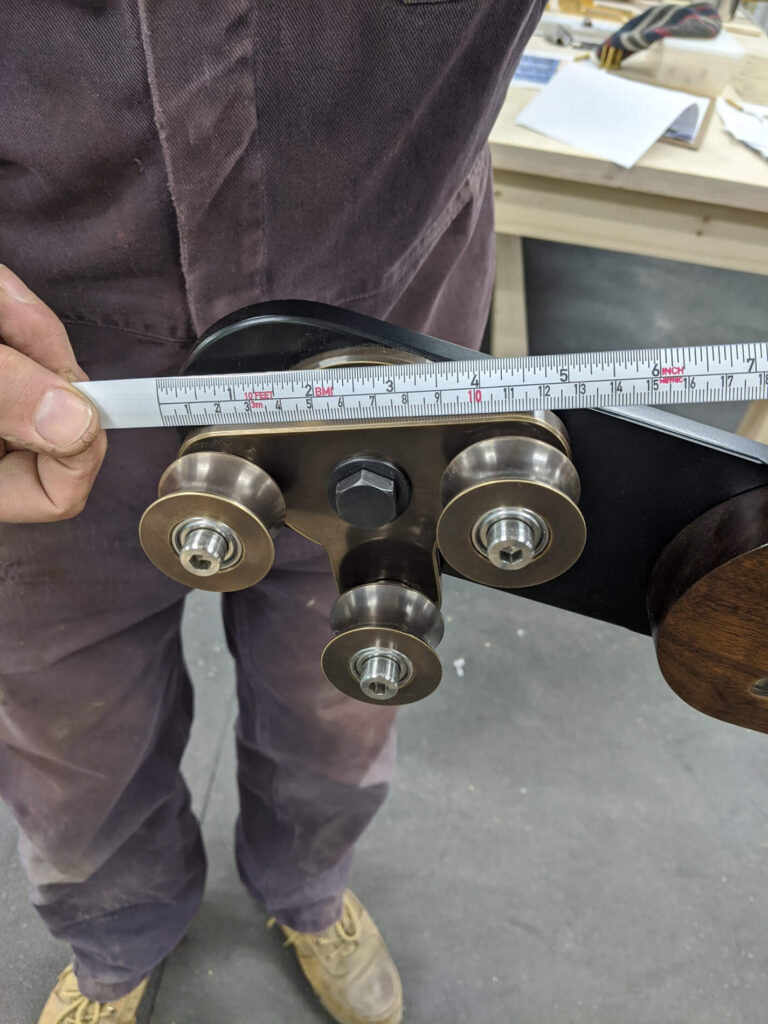 A person in work clothes holds a metal roller bracket whilst measuring it with a ruler in a workshop setting.