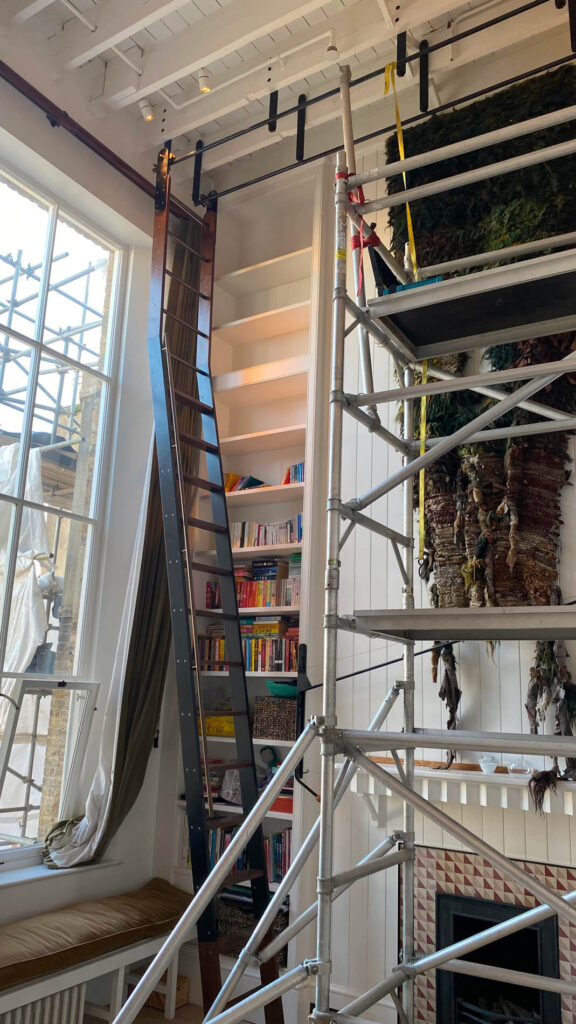 A tall bookcase filled with colourful books stands beside a large window. A rolling ladder leans against the shelf, and metal scaffolding is set up near the fireplace under a high, white ceiling.
