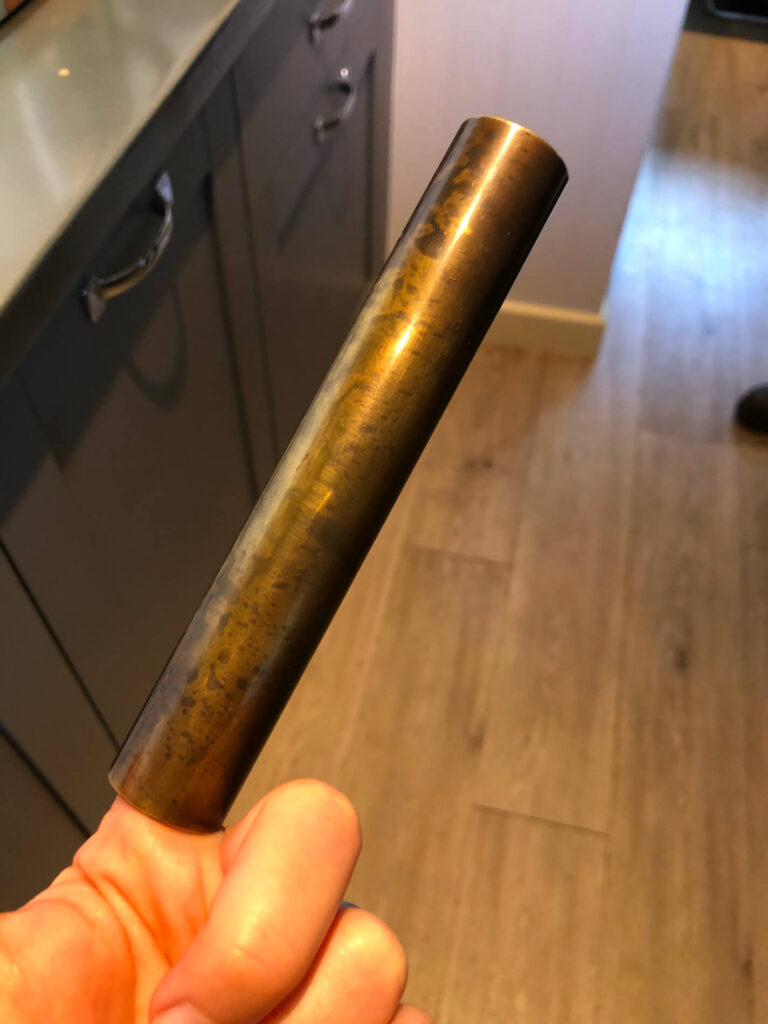 A hand holds a cylindrical metal rod, slightly tarnished, in a kitchen with wooden floors and grey cupboards in the background.