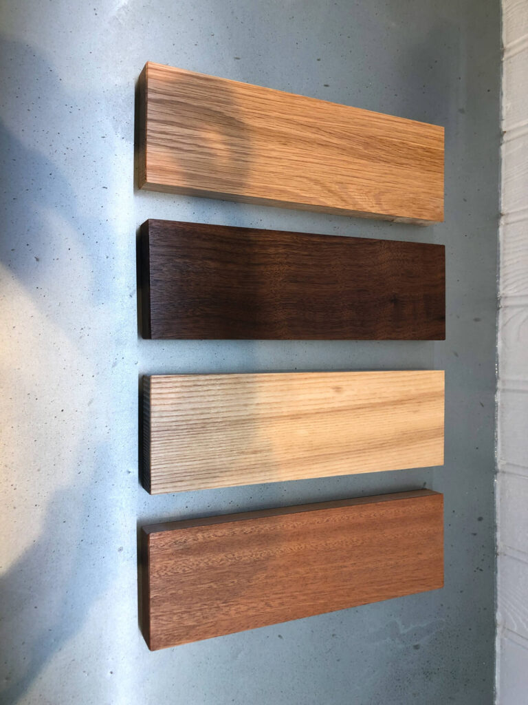 Four rectangular wooden planks of different wood tones are laid out vertically on a grey surface, each with distinct grain patterns and shades ranging from light to dark.