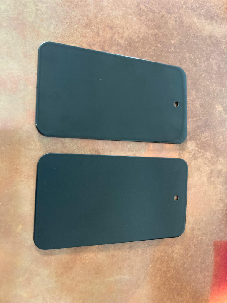 Two rectangular black plastic tags with rounded corners and a small hole in one corner sit side by side on a brown textured surface.