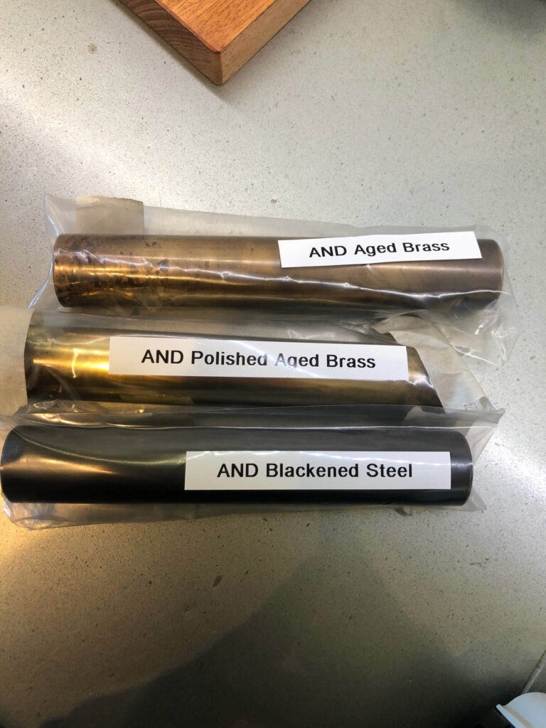 Three labelled metal rods in plastic bags on a counter: “AND Aged Brass” (top), “AND Polished Aged Brass” (middle), and “AND Blackened Steel” (bottom).