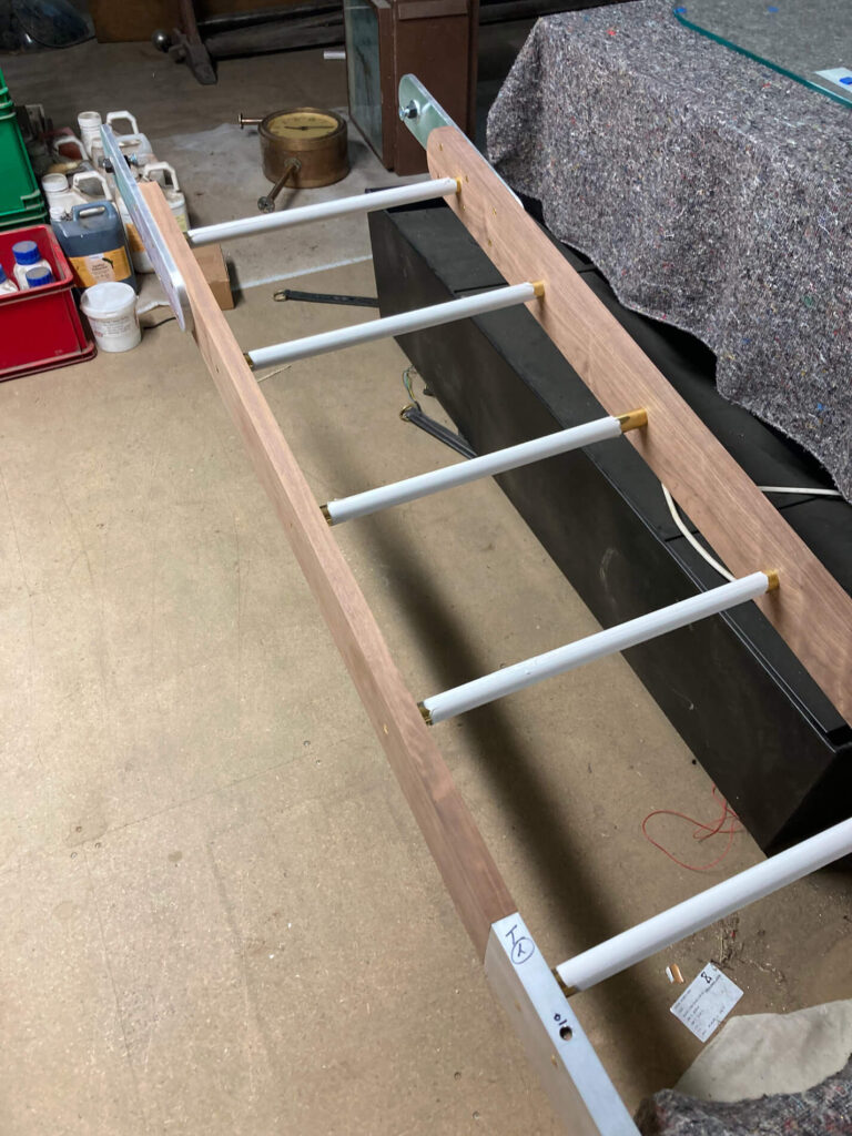 A wooden ladder with six evenly spaced rungs is laid flat on a workshop floor surrounded by tools, containers, and equipment. The setup appears to be a construction or assembly project in progress.