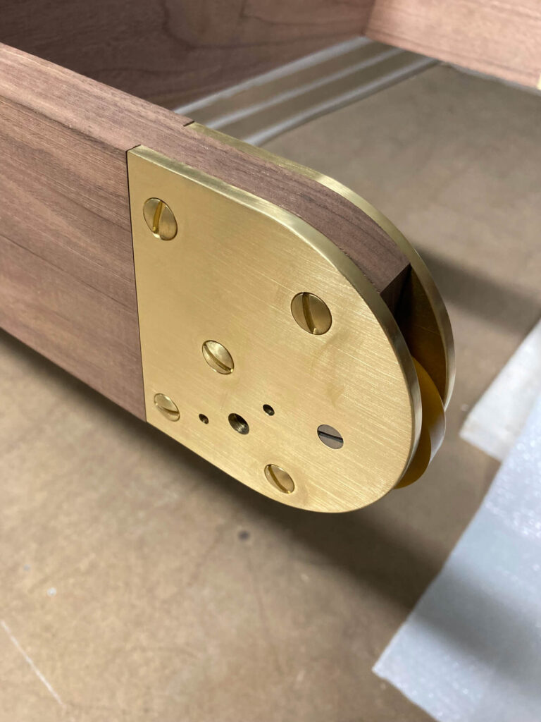 Close-up of a polished brass rolling ladder foot attached to the corner of a wooden library ladder with visible screw holes and smooth, rounded edges.