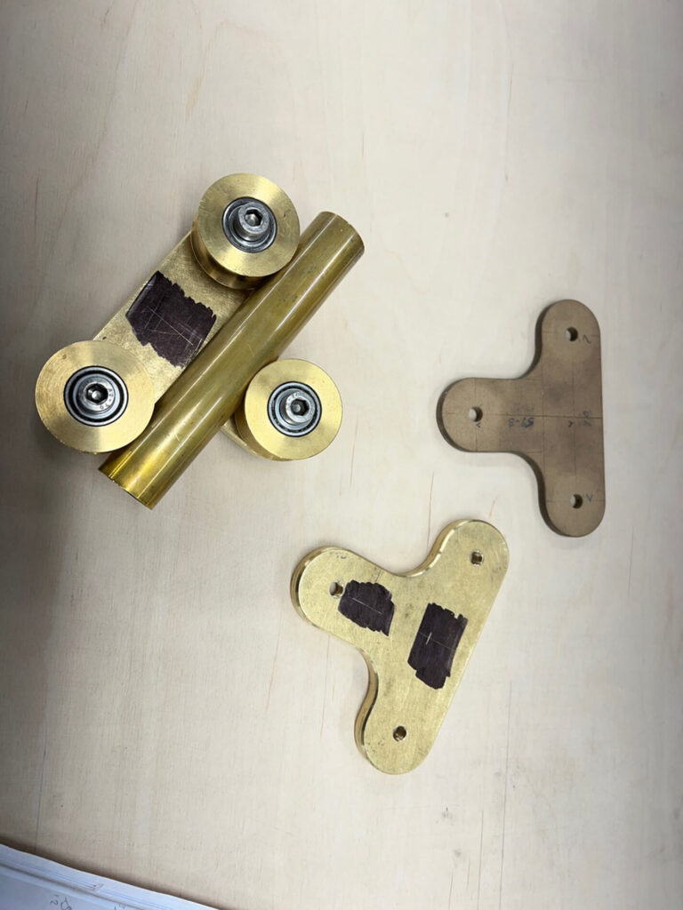 Three brass and metal mechanical parts and two similarly shaped brown templates are placed on a light-coloured wooden surface. The brass parts have attached ball bearings and rectangular shaded areas.