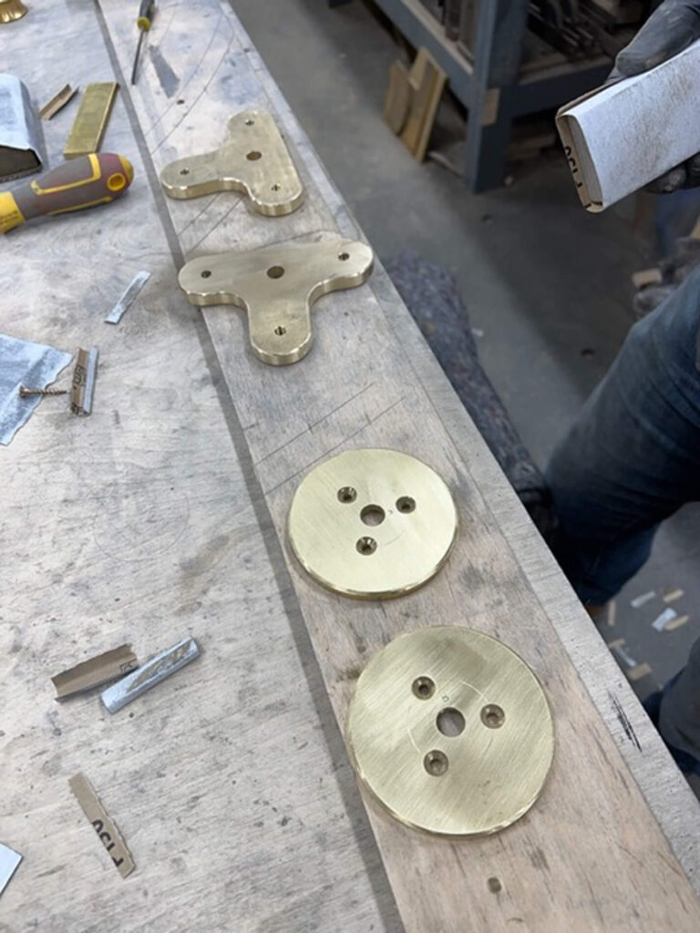 Two brass plates and two brass T-shaped brackets with holes are laid out on a workbench, surrounded by metal swarf and tools. A person’s gloved hand holds a sanding block at the right edge of the image.