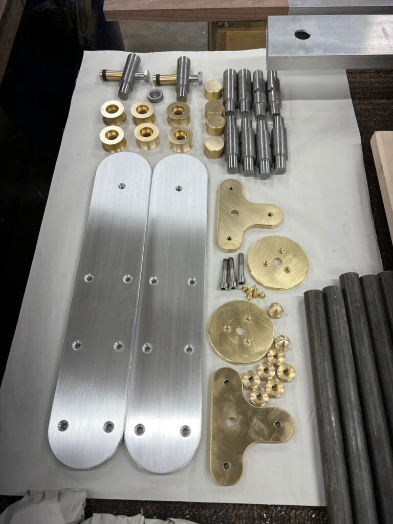 A neatly arranged collection of machined metal parts, including silver and brass plates, gears, rods, cylindrical pins, and small screws, all laid out on a white surface.
