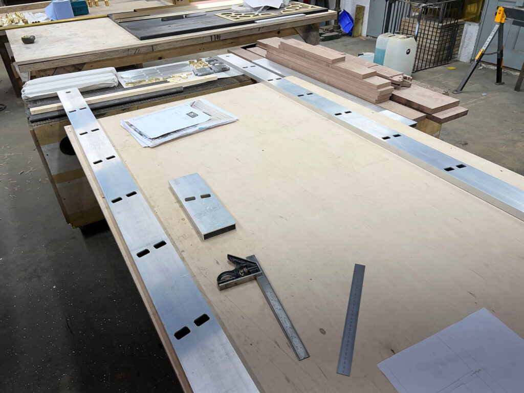 A woodworking bench with metal rulers, a combination square, plans, wooden planks, and other tools and materials laid out in a workshop setting.