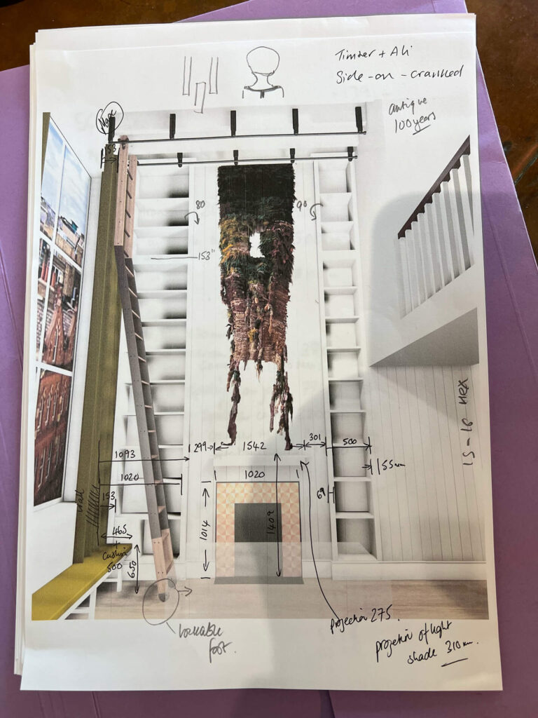 A printed interior design plan with a large vertical artwork above a fireplace, measurements and notes handwritten on the page, and a tall ladder on the left. A staircase and bannister are visible on the right side.