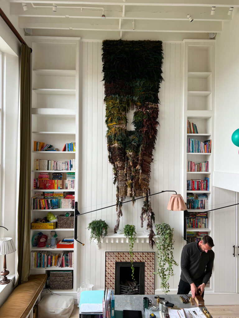 A craftsman stands at a desk in a bright room with tall white walls, built-in bookshelves, a fireplace, hanging plants, and a large textured wall tapestry above the mantelpiece.