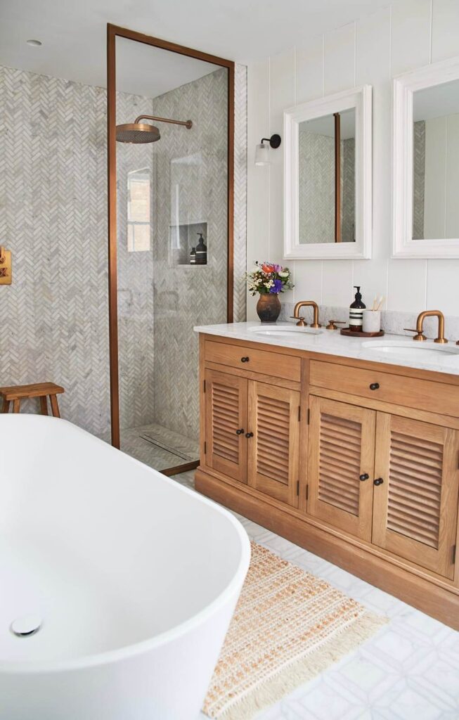 A modern bathroom with a wooden vanity unit, double basins, two mirrors, a white freestanding bath, a patterned rug, and a glass shower with herringbone tiled walls. A vase of flowers sits on the vanity unit.
