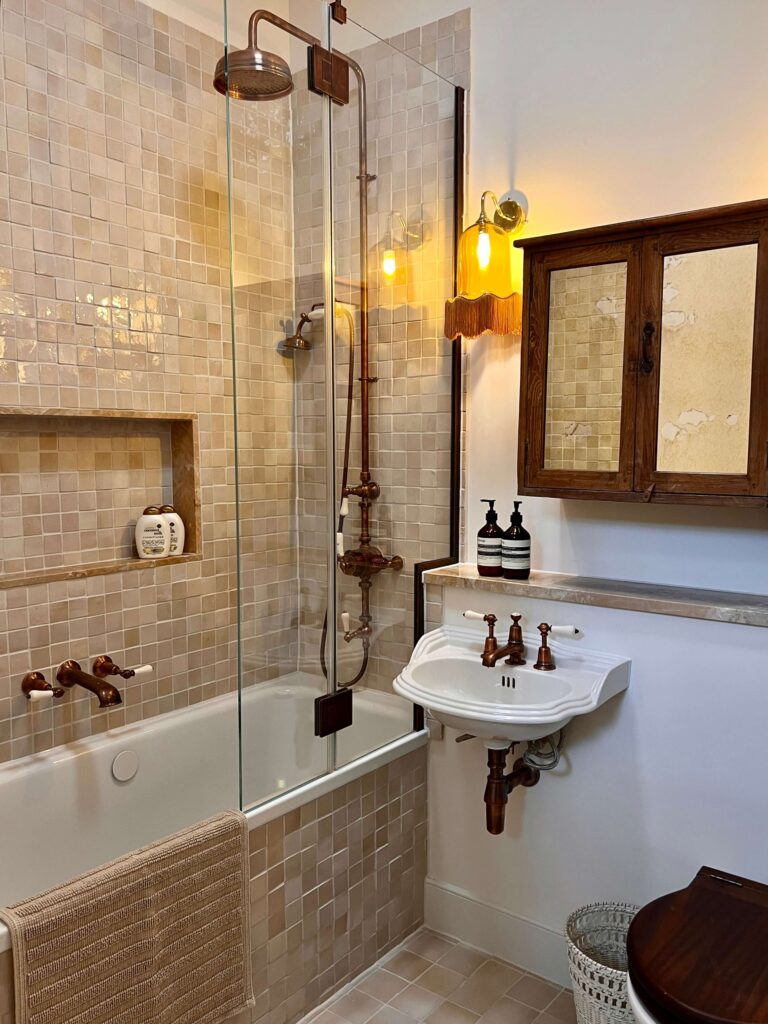 A bathroom with beige square tiles, a bath with glass panel, copper fittings, a white basin, a wooden medicine cabinet, two bottles on the basin, a wall lamp, and a towel hanging on the bath.