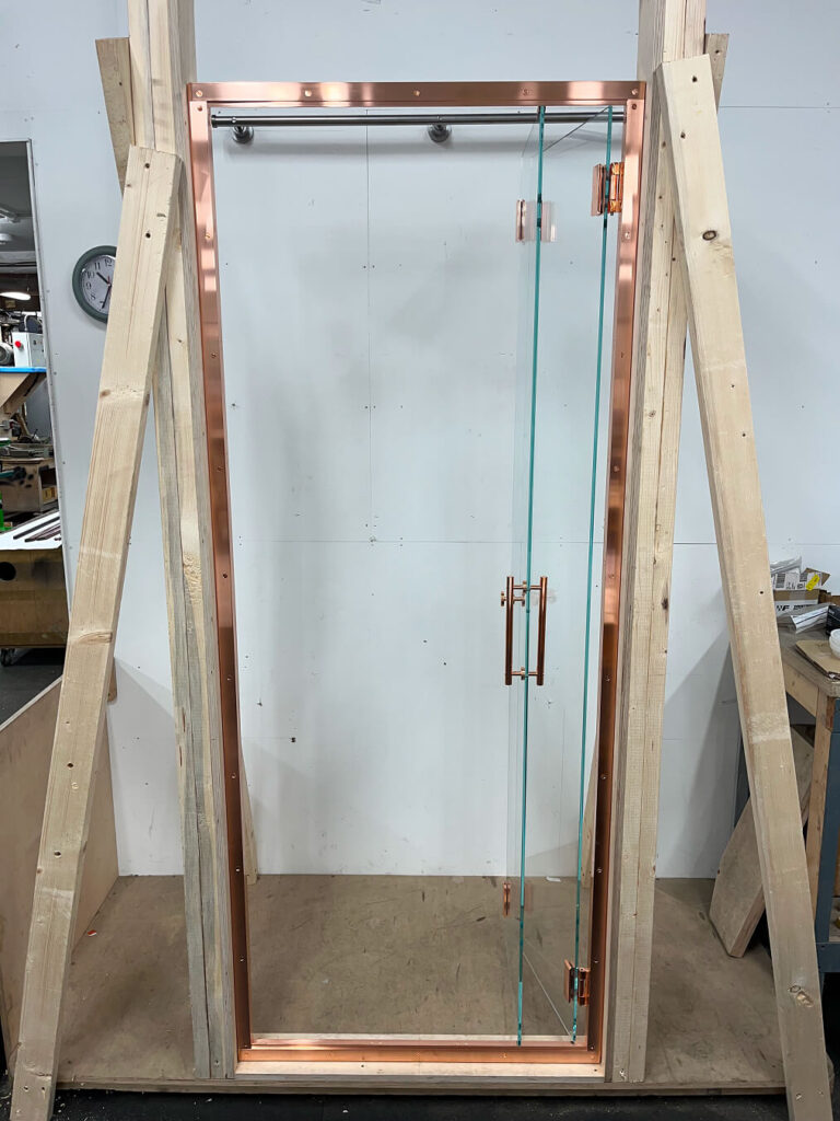 A frameless glass door with a copper frame and handle is mounted in a wooden support structure inside a workshop. The background includes white walls and some visible tools and materials.