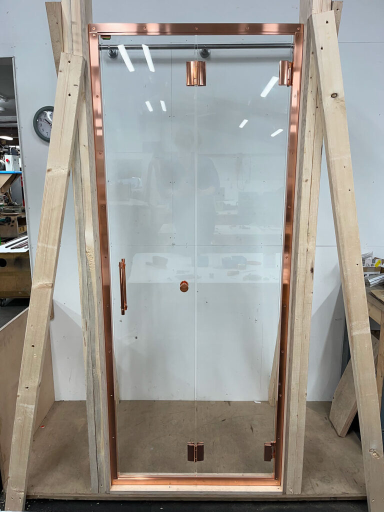 A glass door with a copper frame and handle is mounted in a wooden support structure, set in a workshop with tools, supplies, and a clock visible in the background.