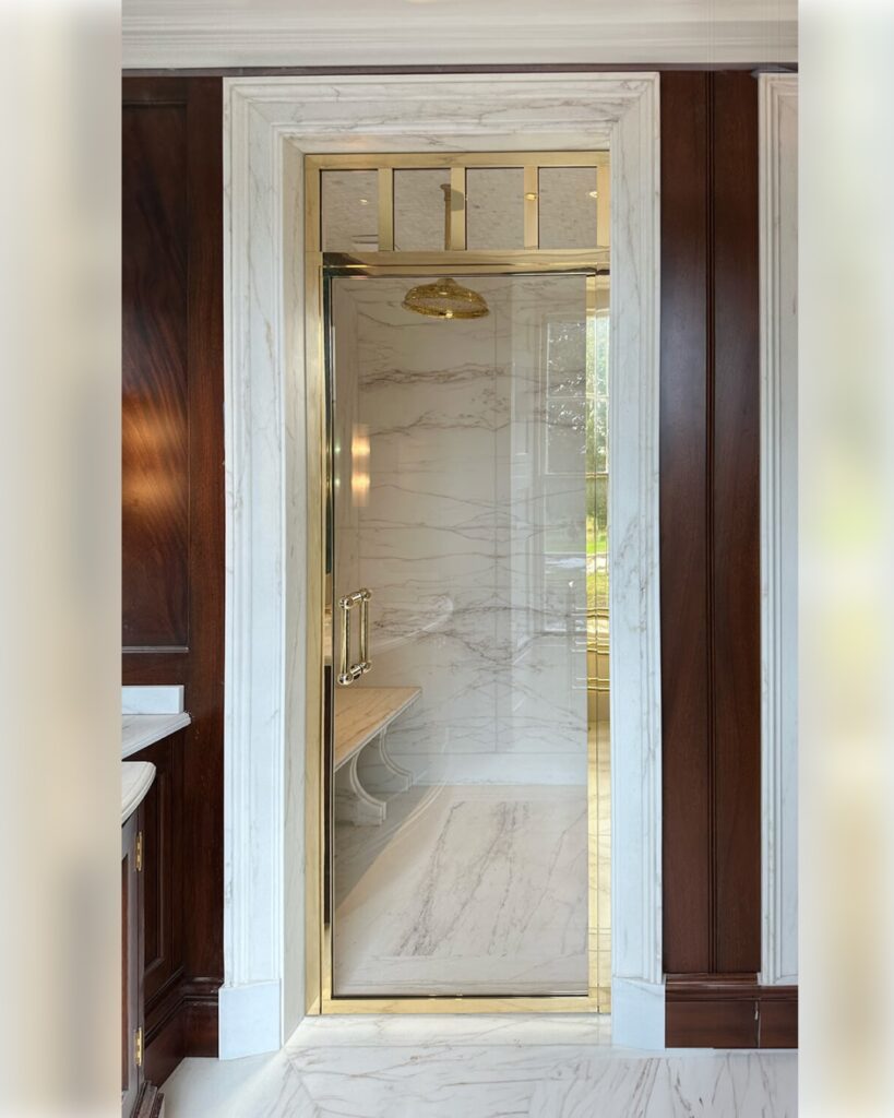 A luxurious shower with marble walls and floors, a glass door framed in gold, a gold showerhead, a marble bench inside, and dark wood panelling on the exterior walls.