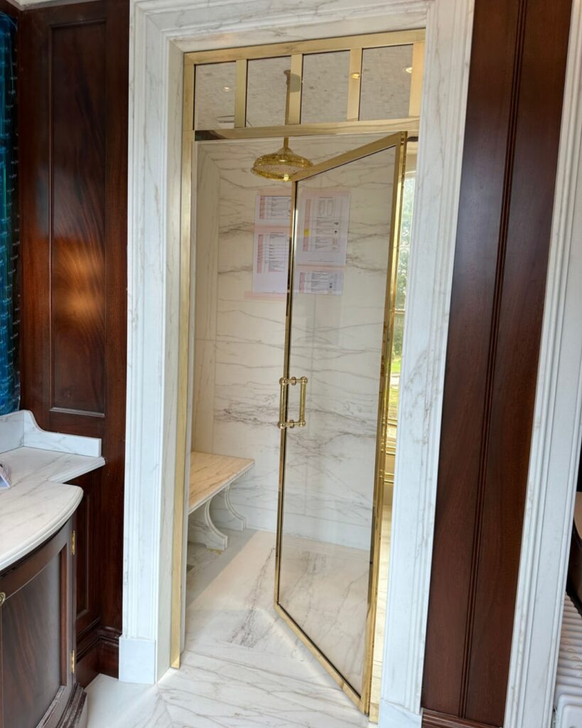 A luxurious shower with gold-framed glass door, gold showerhead, and marble walls and floor. Dark wood panelling surrounds the entrance; a white marble counter is visible on the left. Papers are posted on the back shower wall.