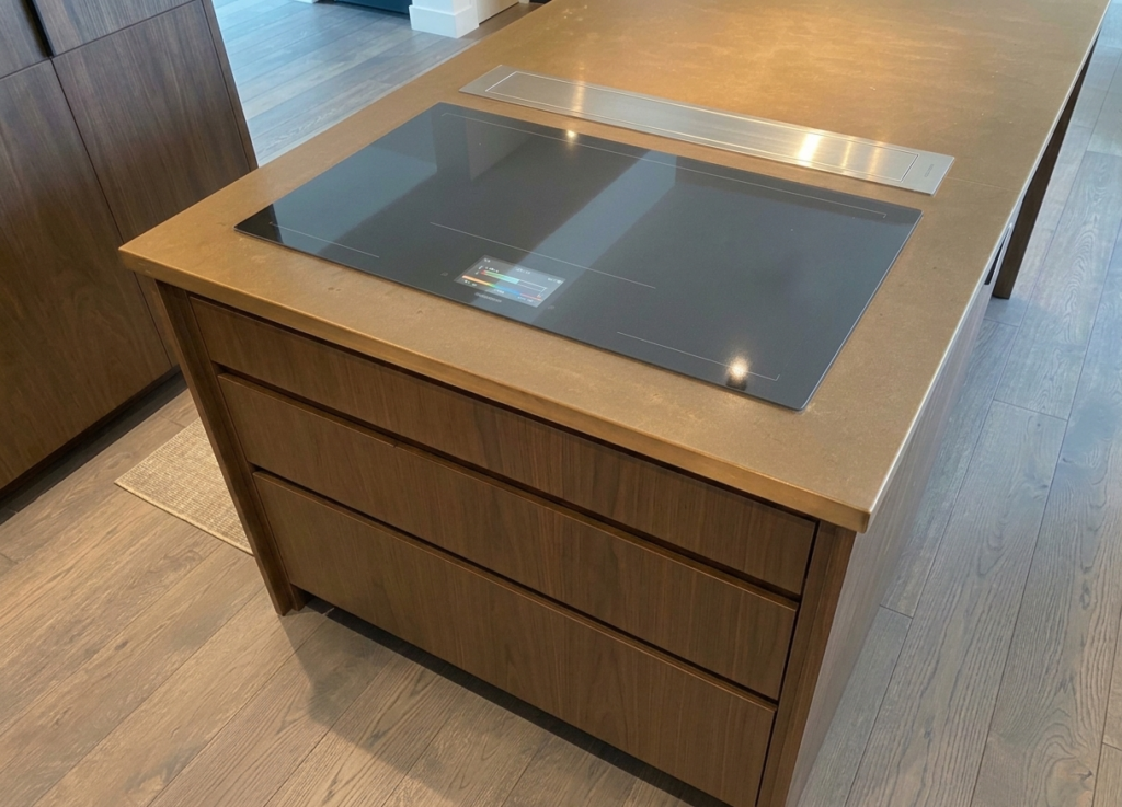 A modern kitchen island with a built-in black glass hob and three wooden drawers beneath, set on a wooden floor. The worktop is smooth and made of copper, and the design is sleek and minimalist. This is one of several matching aged copper kitchen worktops.