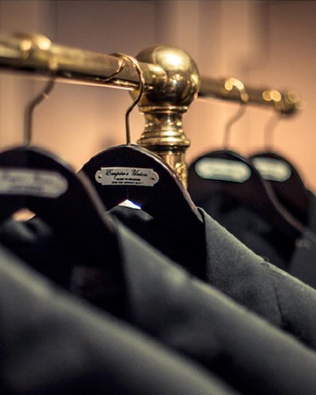 Close-up of several black suits on hangers, neatly arranged on bespoke clothes rail. The focus is on the nearest hanger, while the others gradually blur into the background.