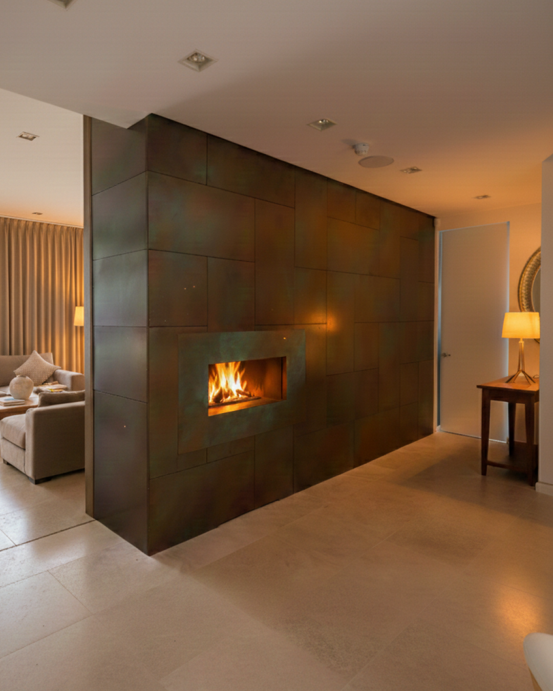 A modern living space featuring a sleek, built-in fireplace set in a copper-panelled wall, with warm ambient lighting, a small side table, and a comfortable seating area with neutral decor.