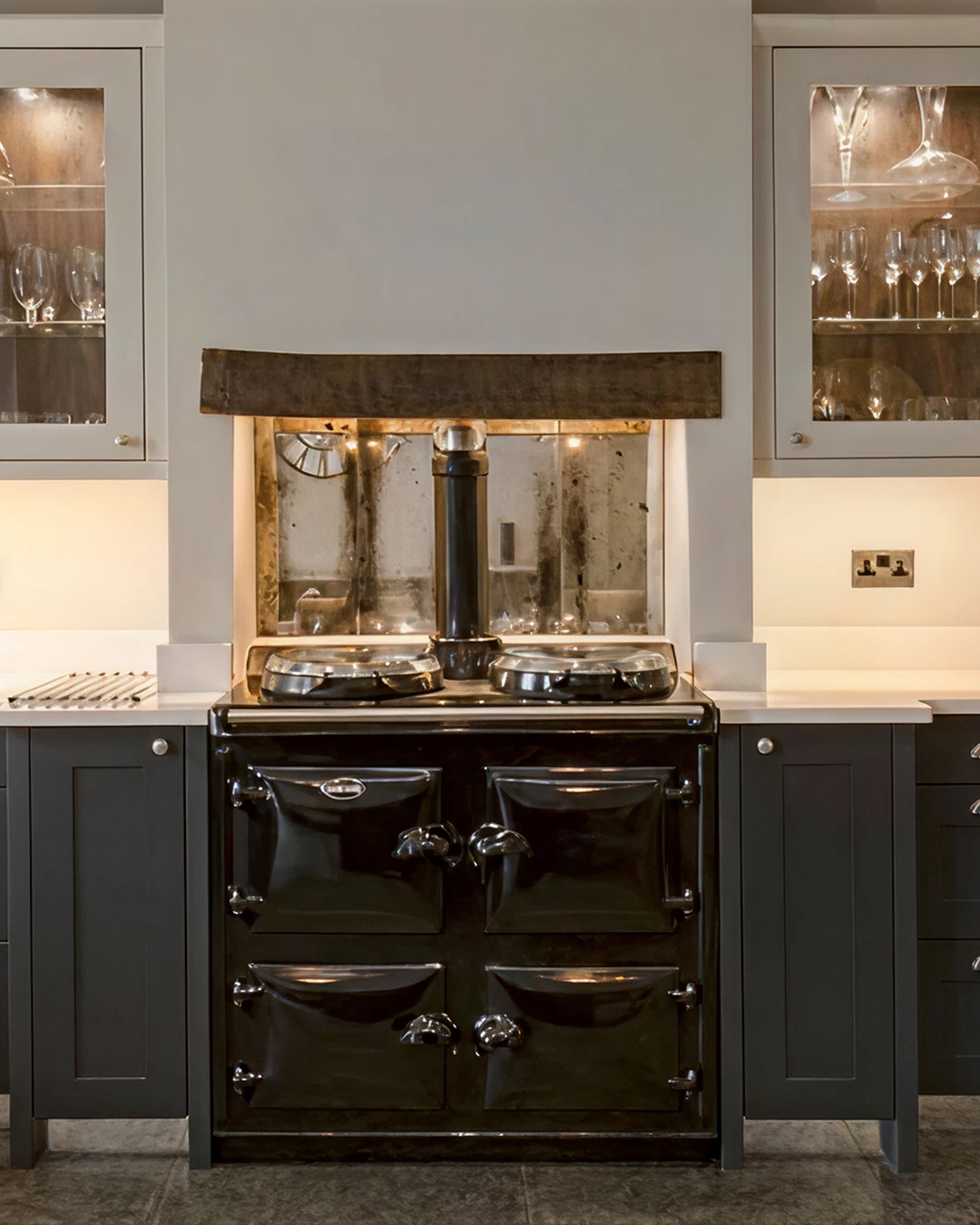 A kitchen hob with a vintage black range cooker beneath a rustic wooden mantel, flanked by grey cabinets and glass-fronted cupboards displaying glassware, reflects softly in bespoke mirrors for added charm.