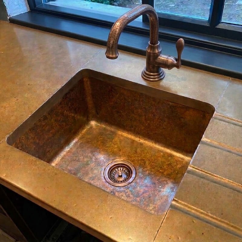 A kitchen sink and worktop made of copper surfaces with a matching tap, situated below a window. The warm, metallic finish and subtle patina add character, while grooves for drainage are neatly positioned on the right side of the sink.