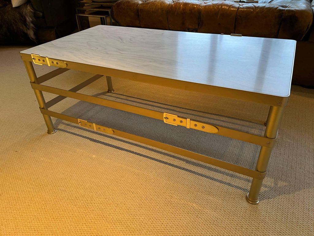 A rectangular coffee table with a light marble effect top and a brass metal frame stands on a beige carpet. The table has two horizontal support bars with decorative brackets and is placed in front of a brown leather sofa.