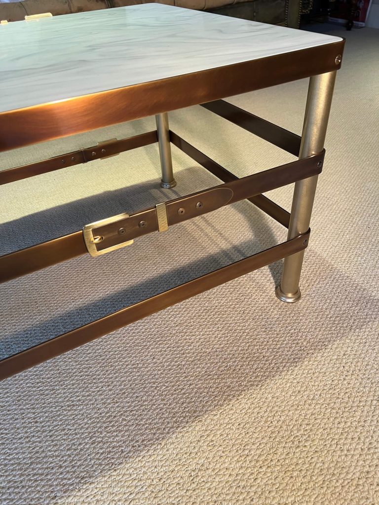 A close-up of a coffee table corner with a metal frame featuring a unique belt buckle design on its horizontal support, standing on a beige carpeted floor. The table top appears to have a light, marble-like surface.