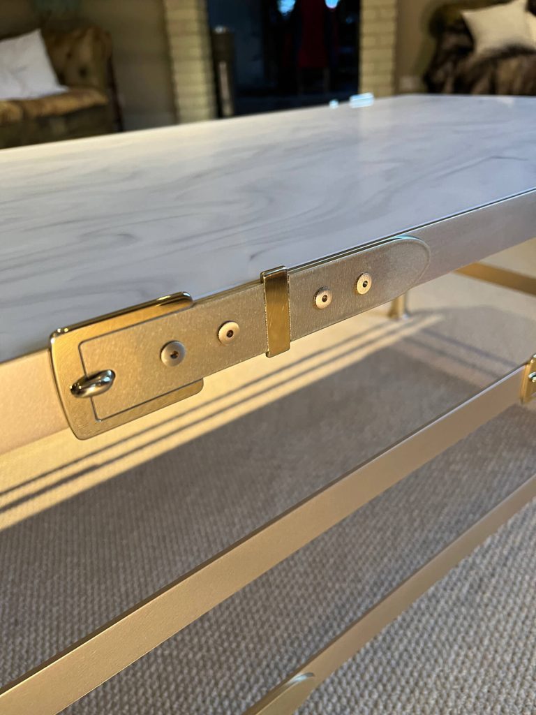 A close-up of a zinc metal belt buckle attached to the underside of a coffee table, with sunlight casting shadows on a textured carpeted floor.