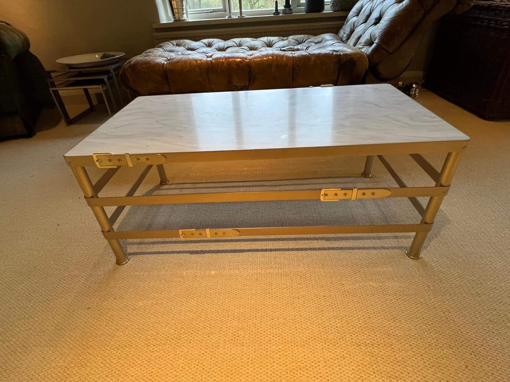A rectangular coffee table with a light-coloured, marble-patterned top and a metallic frame stands on a beige carpet in a cosy living room with a tufted ottoman and window in the background.