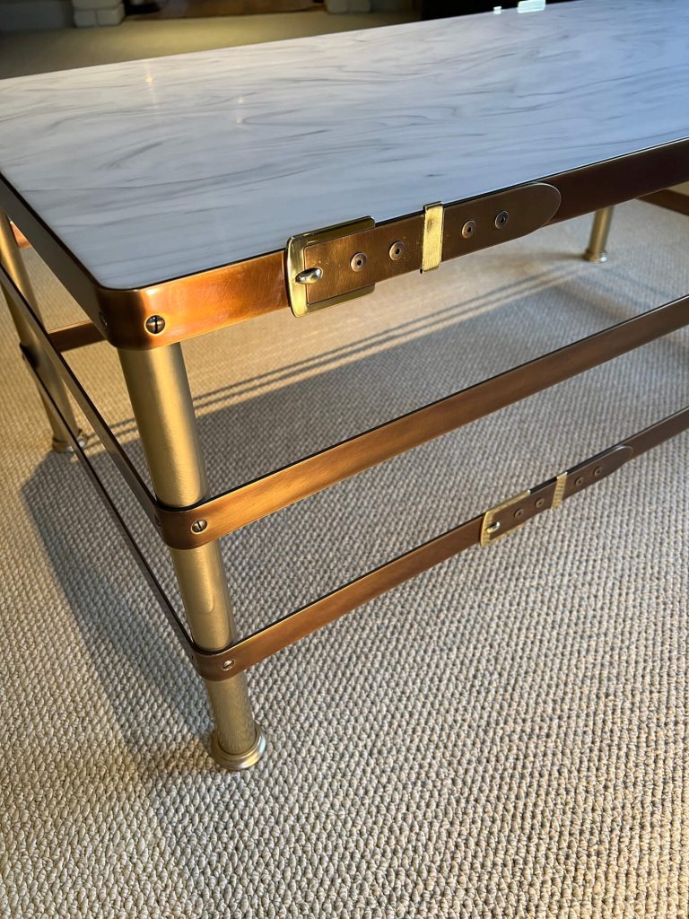 The image shows the corner of a modern coffee table with a white, marbled top and gold-coloured metal legs and trim, featuring brass clasps and fittings, set on a textured beige carpet.