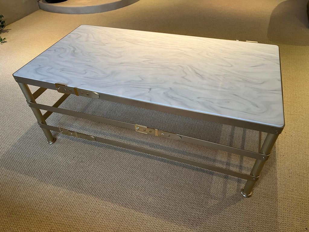 A rectangular coffee table with a marbled light grey top and metallic zinc frame stands on a beige carpeted floor in a well-lit room.