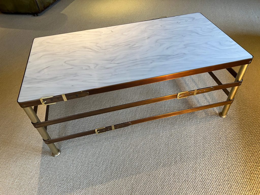 A rectangular coffee table with a white, marble-patterned top and a gold-coloured metal frame stands on a beige carpeted floor. The frame features three horizontal rods with decorative clasp-like accents.