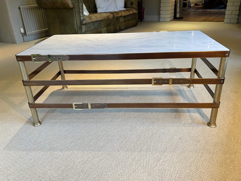 A rectangular coffee table with a white marble top and brass frame, featuring three horizontal bands secured with metallic buckle-like fasteners, placed on a beige carpet in a cosy living room.