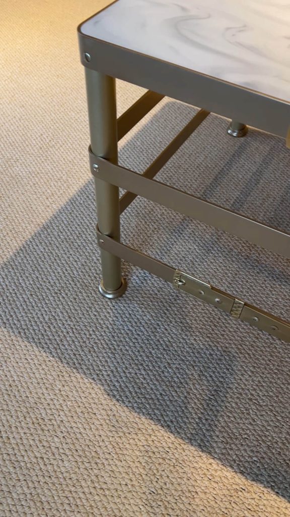 A close-up view of the lower legs and base of a metal coffee table, standing on a textured beige carpet with sunlight creating shadows on the floor.