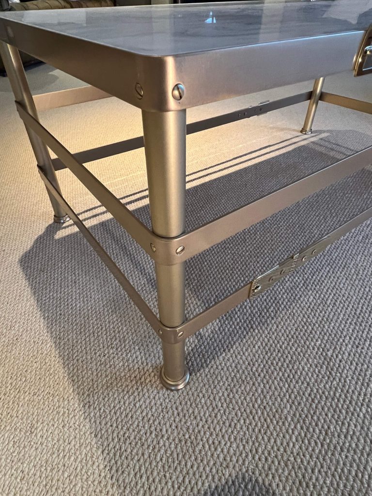 A close-up of a metal-framed coffee table with a marble effect top, set on a beige textured carpet. The frame is silver coloured with visible bolts and a lower support bar.