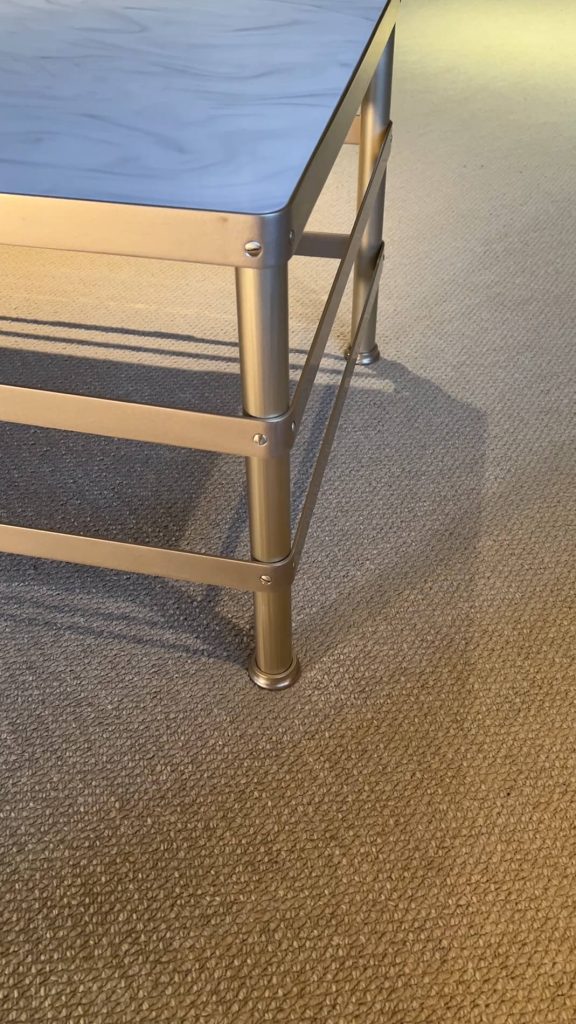 A close-up of the corner of a metal coffee table with a light-coloured top, standing on beige textured carpet. The table legs have bolts and connecting metal bars for support.
