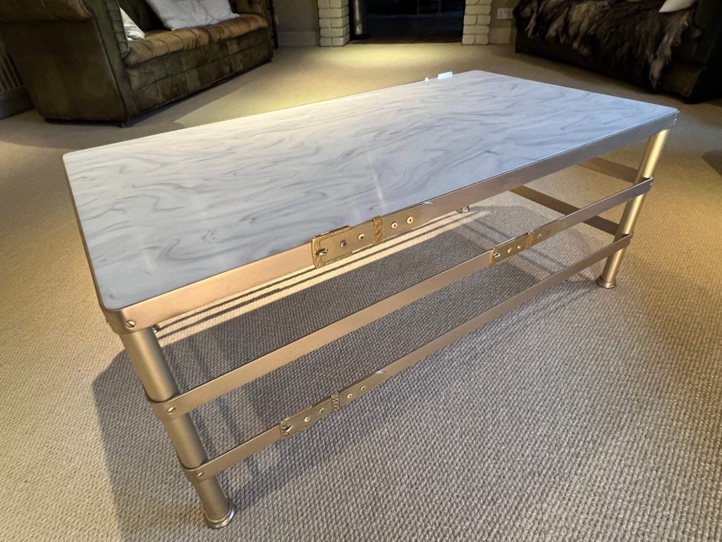 A modern coffee table with a light, marbled surface and a zinc metal frame stands on a beige carpet in a living room with sofas and a fireplace in the background.