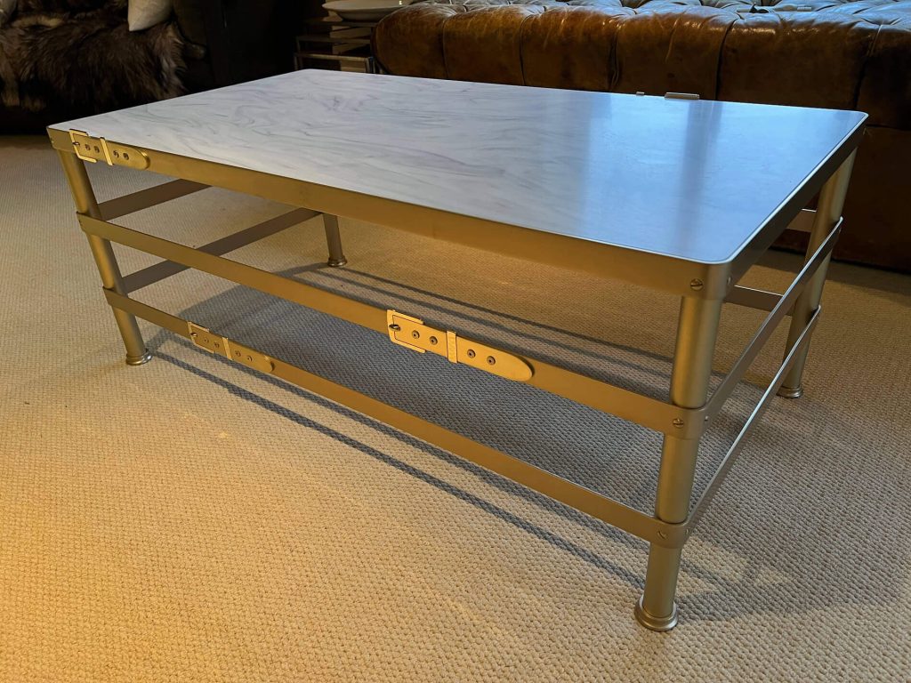 A modern rectangular coffee table with a light-coloured, smooth wooden top and a metallic frame featuring horizontal bars and exposed hinges, standing on a beige carpet in a lounge.
