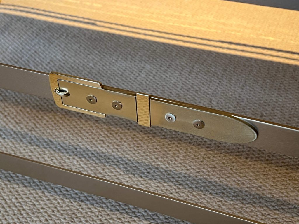 Close-up of a zinc buckle fastening two brown metal bars together, placed on a textured beige woven surface with sunlight casting shadows.