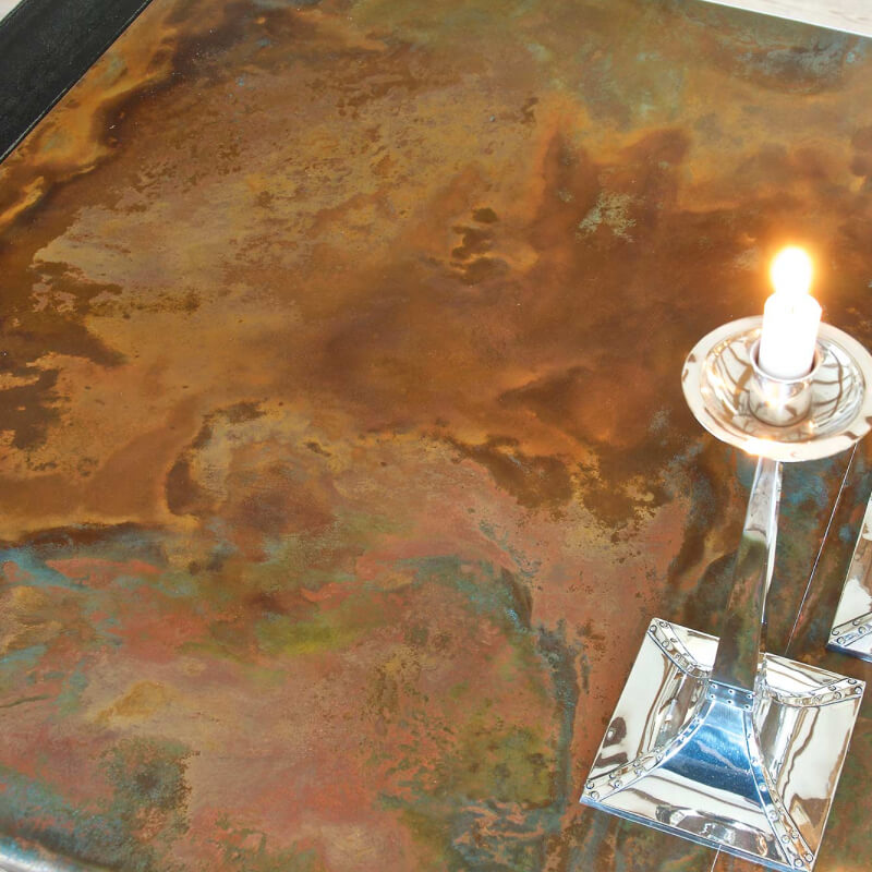 A single lit candle in a silver holder sits on a surface with a textured, rust-coloured, and multicoloured patina, reminiscent of aged copper surfaces. The background appears metallic with earthy tones and an abstract pattern.
