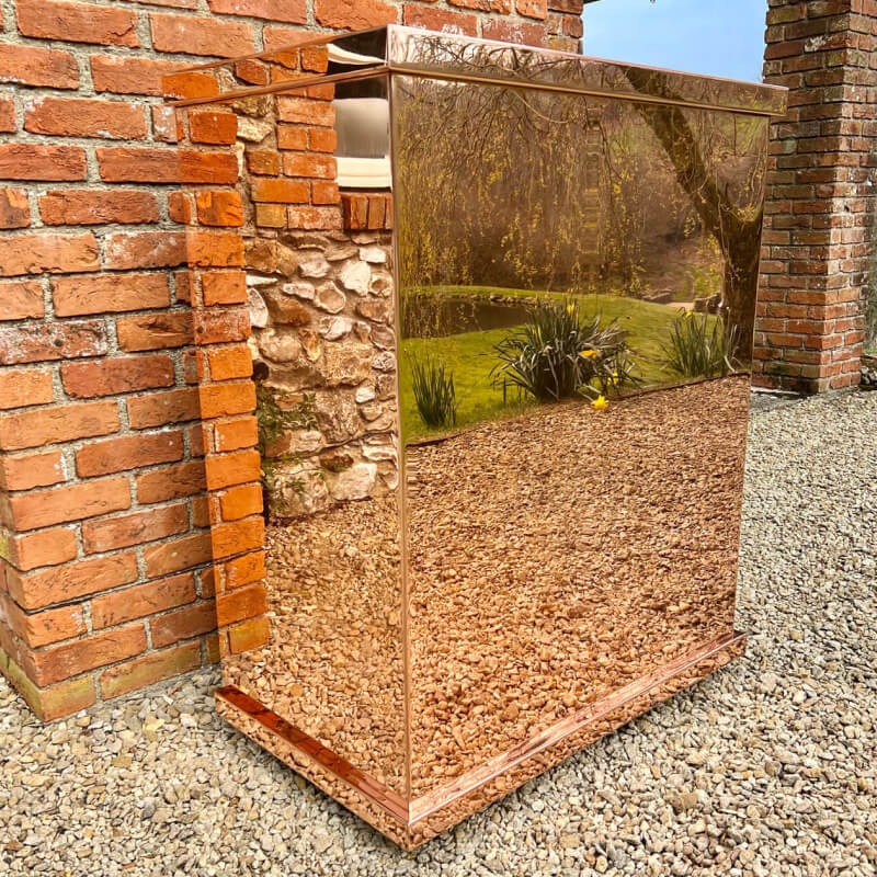 A shiny, copper coloured mirrored cabinet with copper surfaces stands on gravel beside a brick and stone wall, reflecting the surrounding garden and landscape in its surface.