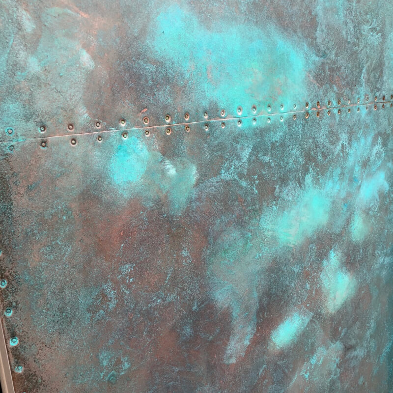 Close-up of a weathered metal surface with a row of rivets. The mix of rusty brown and turquoise-green patina evokes aged copper surfaces, creating a richly textured appearance.