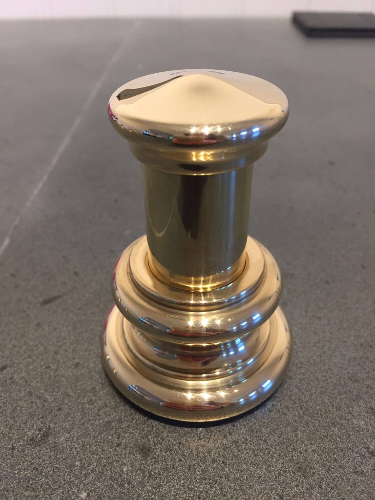 A shiny brass fixed valet hook with a rounded top and stacked circular base, sits on a grey surface.